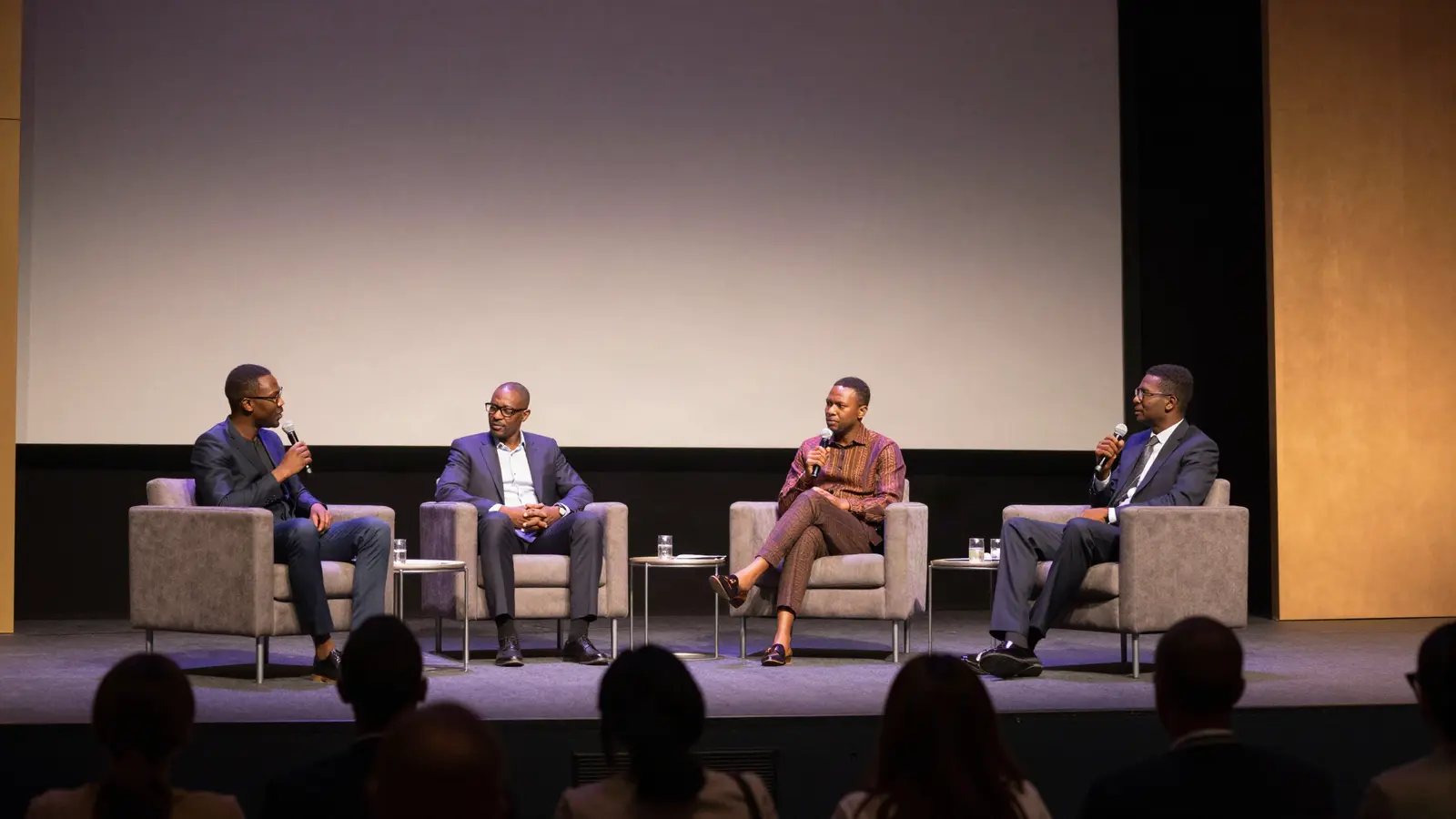 AI-generated illustration: four speakers in armchairs on a stage, one speaking into a microphone, with the audience in silhouette